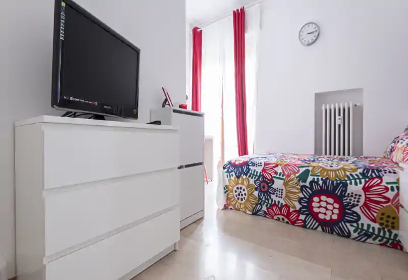 Camera da letto luminosa e colorata con copriletto floreale, mobili bianchi e ampia finestra con tende rosse che lasciano entrare luce naturale.