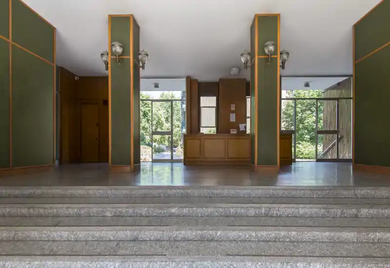 Ingresso ampio e simmetrico con banco reception, gradini in pietra lucida e grandi finestre che lasciano entrare luce naturale.