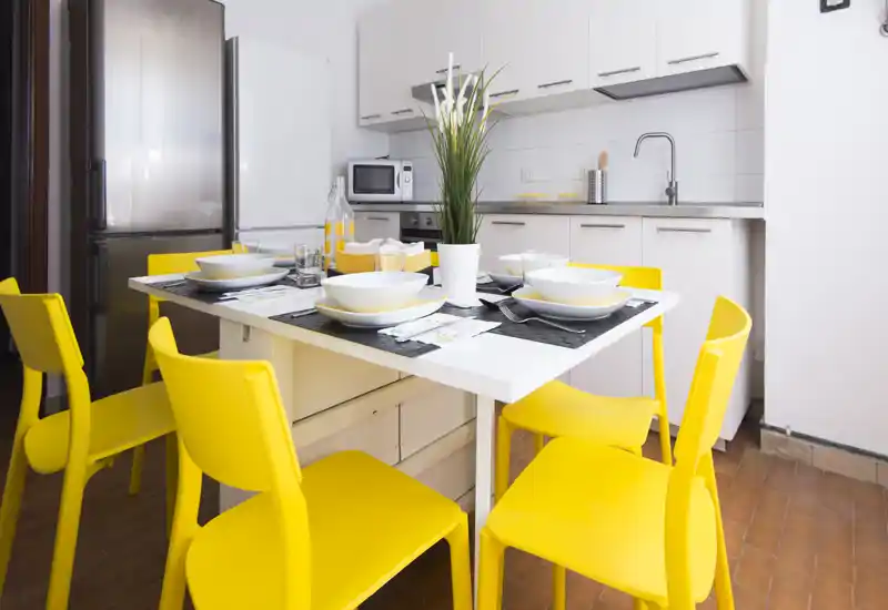 Cucina moderna e luminosa con tavolo apparecchiato e sedie giallo vivace. Layout compatto con mobili bianchi, frigorifero in acciaio e piccoli elettrodomestici, ideale per pranzi informali.