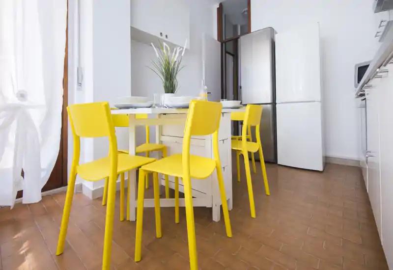 Angolo cucina moderno con un piccolo tavolo da pranzo e quattro sgargianti sedie gialle, mobili bianchi e frigorifero—ben illuminato e allestito per foto di affitto.