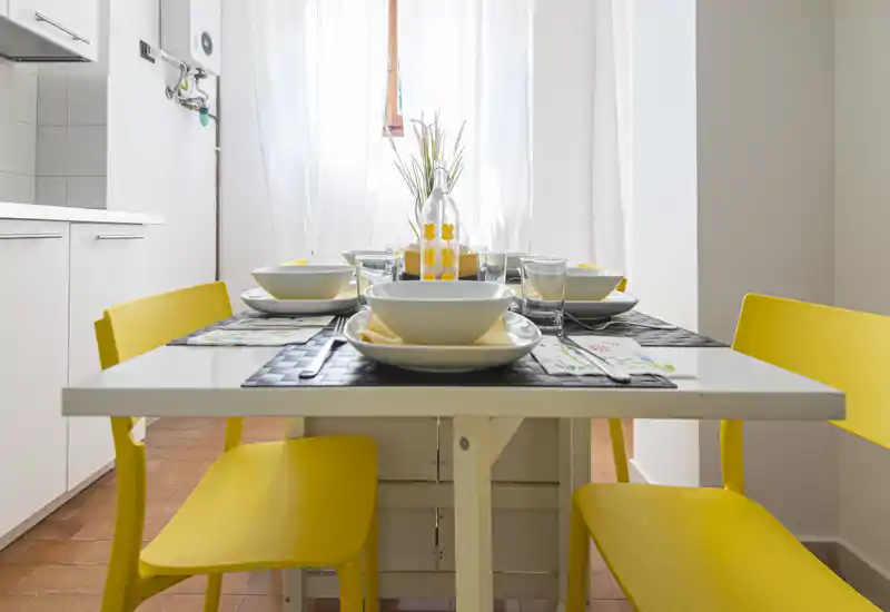 Area pranzo luminosa e ordinata con tavolo bianco e sedie gialle, apparecchiata per un pasto informale vicino alla finestra.