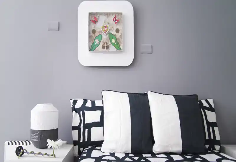 Rincón de dormitorio moderno y luminoso con cojines en blanco y negro y ropa de cama con estampado contra una pared gris suave, con una lámpara de mesita y un cuadro decorativo.
