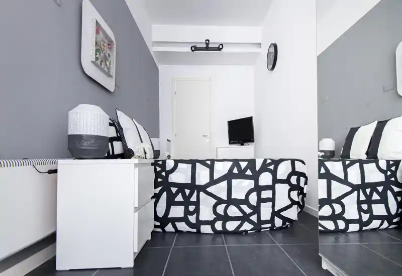 Dormitorio minimalista en blanco y negro con colcha estampada, mesita de noche y cojines decorativos; ambiente luminoso y moderno con un gran espejo que amplía el espacio.