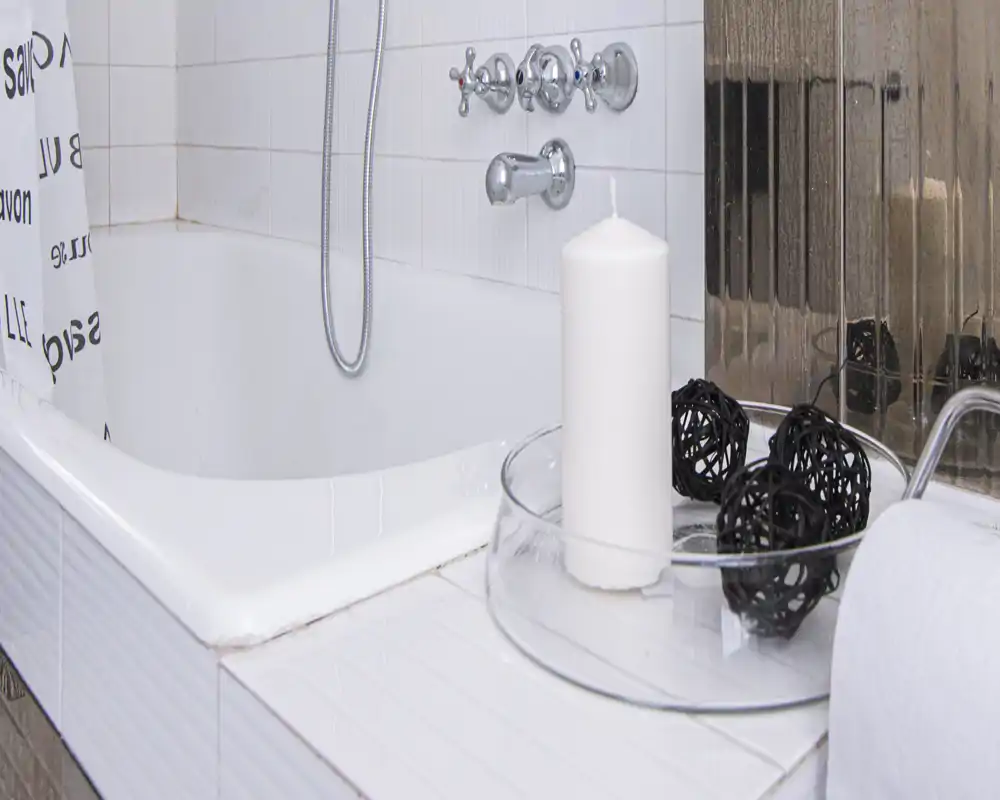 Rincón de baño limpio y luminoso con bañera blanca, grifería cromada, ducha de mano, vela decorativa y cuenco con bolas de rattan.