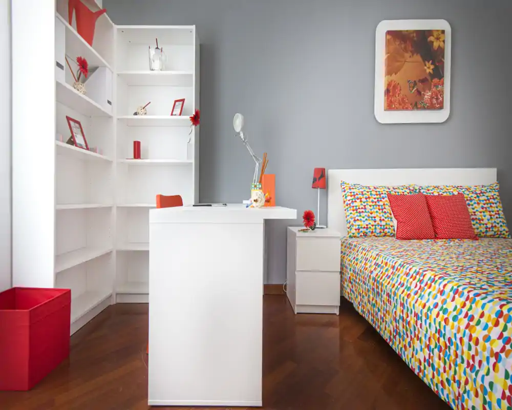 Dormitorio moderno y luminoso con colcha de estampado colorido, dos cojines rojos, mesita blanca y un escritorio ordenado. Suelo de madera cálida y detalles rojos coordinados que crean un ambiente fresco y acogedor.