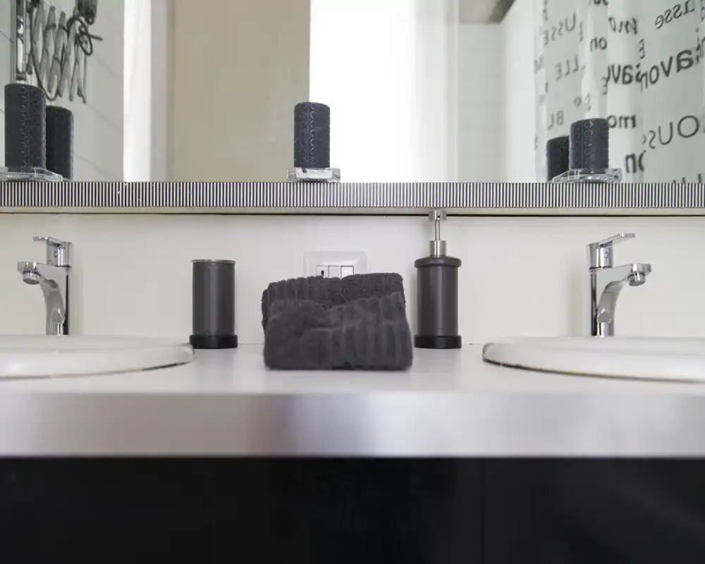 Mueble de baño moderno con dos lavabos, grifos cromados, toalla doblada en el centro y accesorios frente a un gran espejo.