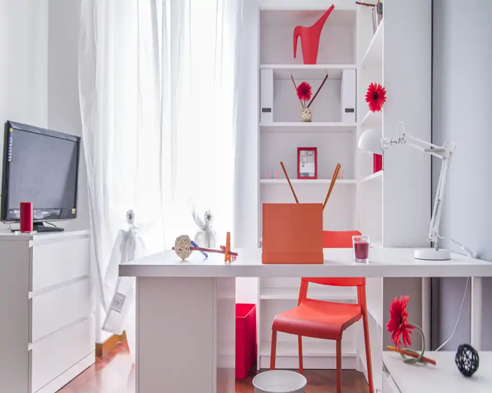 Rincón de salón/oficina moderno y luminoso con muebles blancos y llamativos toques rojos; escritorio ordenado, silla y estantería que crean un espacio de trabajo con estilo.