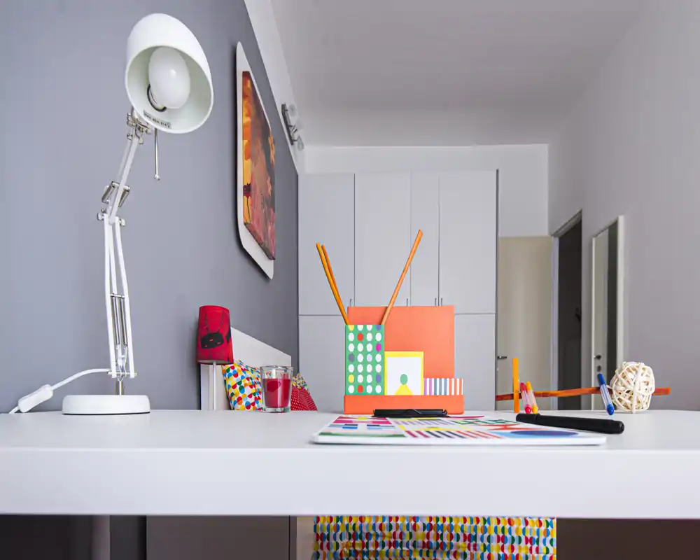 Rincón de dormitorio luminoso y moderno con un escritorio blanco en primer plano, papelería colorida y accesorios, y la cama parcialmente visible detrás. Líneas limpias, acentos vivos y armarios empotrados ofrecen un aspecto funcional y fresco.