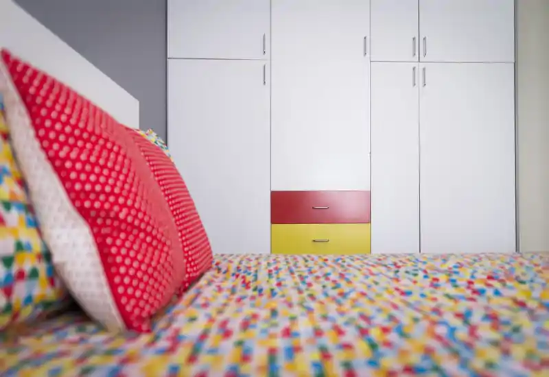 Dormitorio luminoso y colorido en primer plano, ropa de cama estampada y cojines decorativos frente a armarios empotrados blancos.