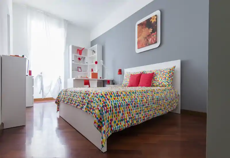 Dormitorio moderno y luminoso con colcha colorida y cojines, cabecero blanco y zona de estudio junto a la ventana—bien iluminado y con buena decoración.