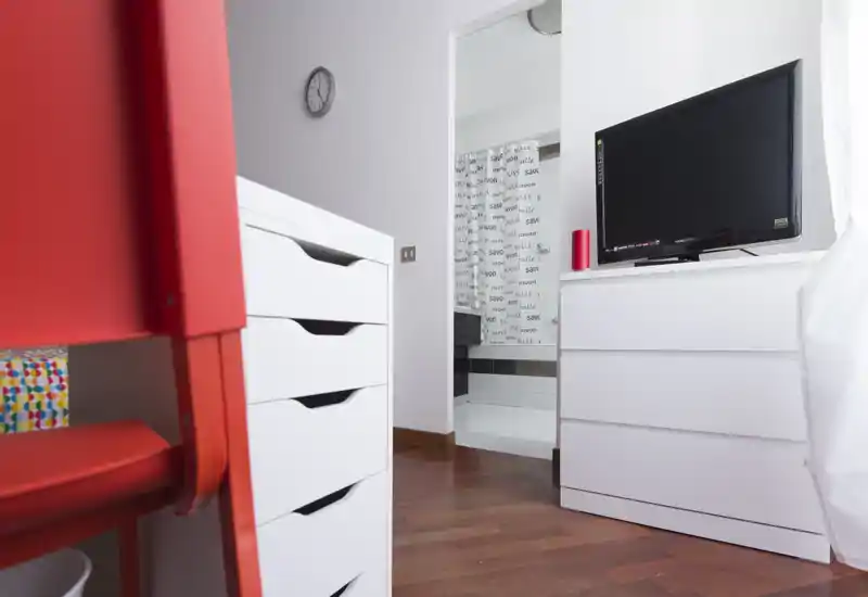 Rincón de dormitorio moderno y compacto con cómodas blancas, televisor y suelo de madera claro; se ve el baño a través de la puerta abierta. El espacio está limpio, bien iluminado por luz natural y amueblado de forma sencilla y contemporánea.