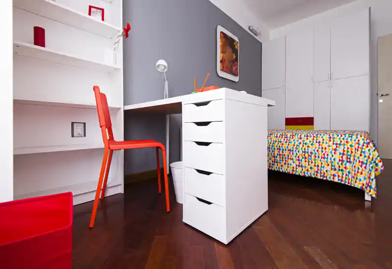 Dormitorio moderno y luminoso con cama individual con ropa de cama colorida, escritorio blanco con cajones y silla roja — ideal para estudiante o ocupante individual.