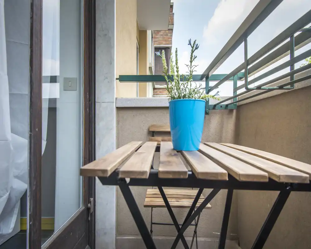 Pequeño balcón amueblado con mesa y silla plegables de madera y una maceta azul con planta, ideal para el café matutino.