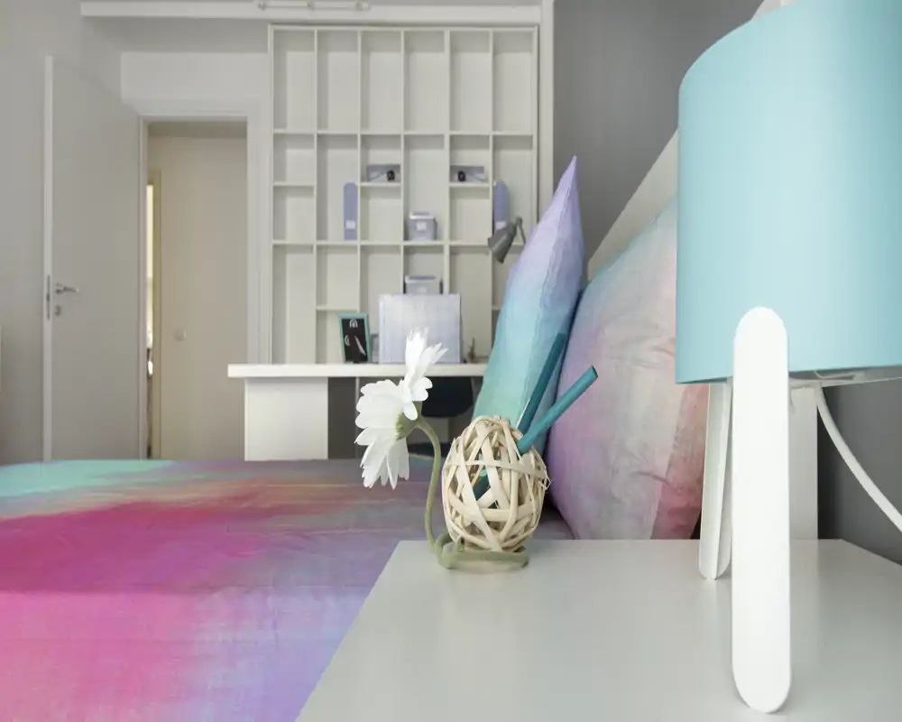 Dormitorio moderno y luminoso con edredón colorido, cojines decorativos y una lámpara de mesita; se ven estanterías ordenadas y un pequeño escritorio al fondo.