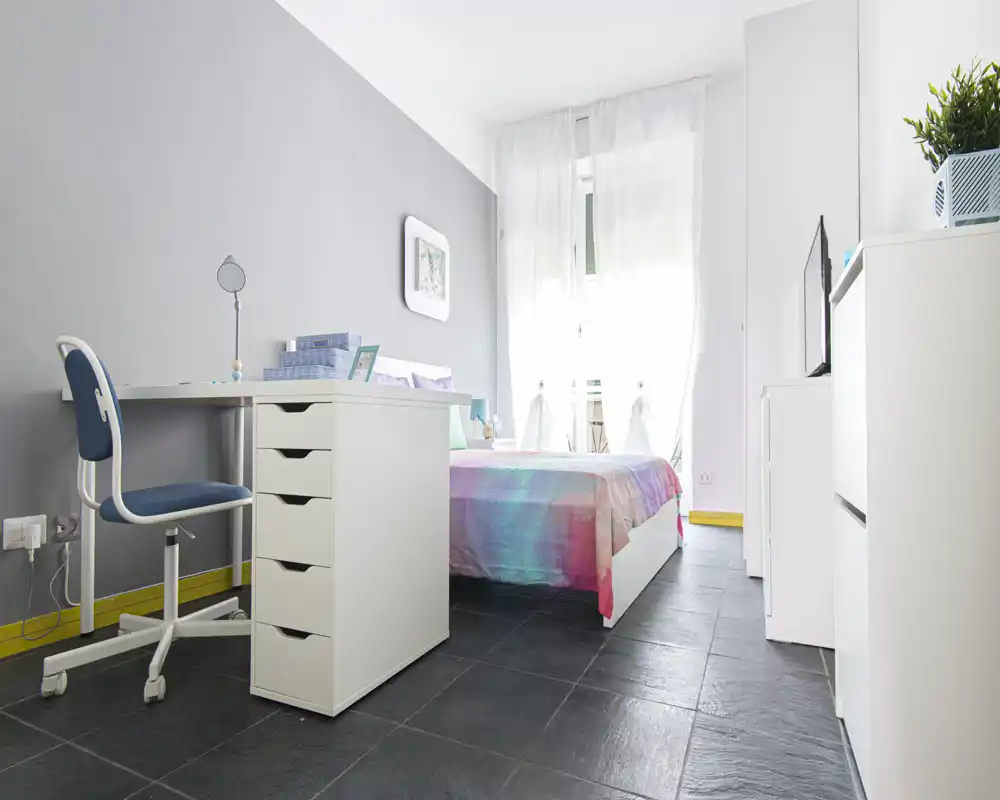 Dormitorio moderno y luminoso con colcha colorida, zona de escritorio compacta y ventana grande que aporta mucha luz natural. Líneas limpias y decoración minimalista crean un ambiente fresco y acogedor, perfecto para un estudiante o una persona sola.
