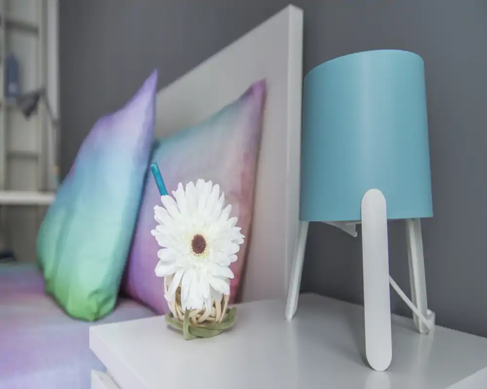Primer plano del área de la mesita de noche de un dormitorio con almohadas coloridas, una lámpara turquesa y una flor blanca decorativa sobre la mesita. La composición resalta textiles suaves y un estilo de dormitorio moderno y ordenado.