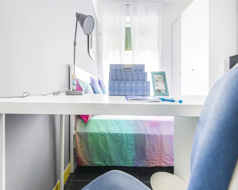 Dormitorio luminoso y compacto con una cama de colores parcialmente visible bajo un escritorio blanco; mobiliario moderno y luz natural que crean un ambiente fresco y ordenado.