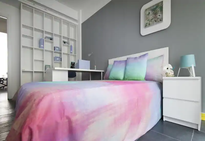 Dormitorio moderno y luminoso con una cama amplia y colorida, mesita de noche y un escritorio ordenado con estanterías — ideal para una estancia cómoda o el teletrabajo.