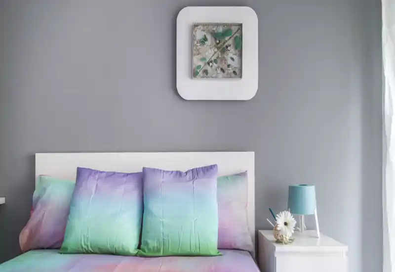 Dormitorio moderno y luminoso con cama de tonos pastel y cojines coloridos; cabecero blanco y mesita con lámpara que crean un ambiente fresco y acogedor.