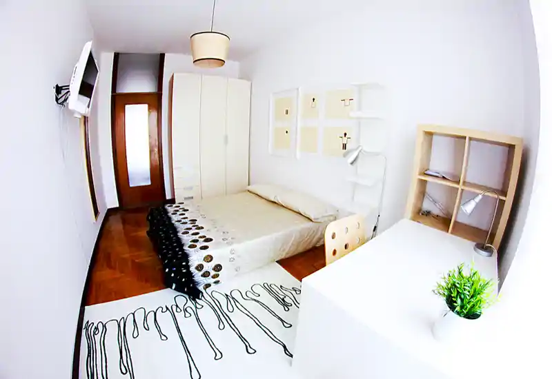 Camera da letto luminosa e moderna con letto matrimoniale, lampada da comodino, armadio e scrivania ordinata. Toni neutri e arredo essenziale creano un ambiente tranquillo e accogliente ideale per soggiorni brevi.