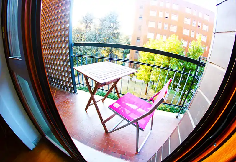 Piccolo balcone soleggiato con un tavolino pieghevole e una sedia pieghevole rosa che affaccia su un cortile verde; ideale per il caffè mattutino.