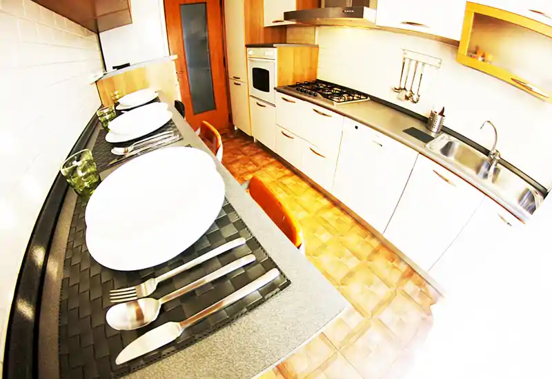 Cucina moderna compatta con lunga superficie, forno incassato, piano cottura a gas e lavello; posti a tavola in stile bar e dettagli in legno caldo rendono l'ambiente accogliente.