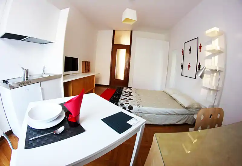 Estudio moderno y luminoso con cama doble, pequeña cocina y mesa; paredes blancas, suelo de madera y decoración minimalista que crean un ambiente acogedor.