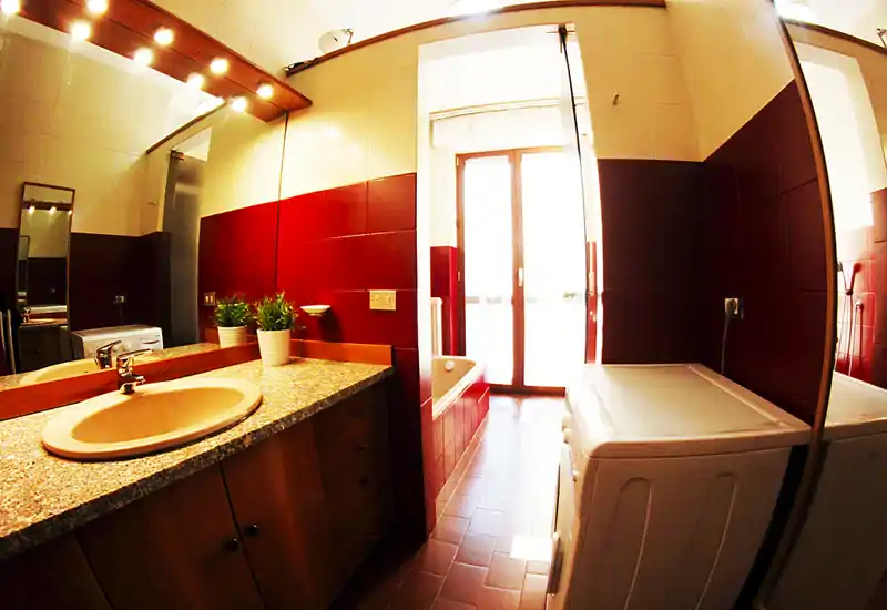 Baño moderno y luminoso con lavabo sobre encimera de piedra, bañera empotrada y lavadora junto a una gran ventana/puerta que aporta abundante luz natural.