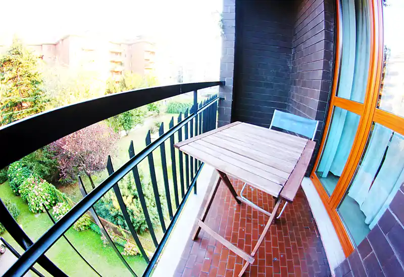 Balcón soleado y estrecho con una pequeña mesa de madera y una silla con vistas a un jardín verde — ideal para el café de la mañana.