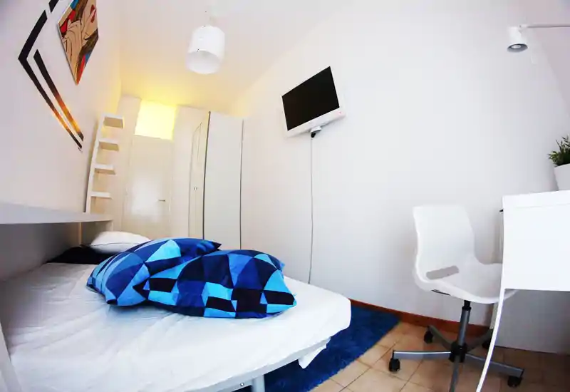 Compact, modern bedroom with a low single bed dressed in geometric blue bedding, a wall-mounted TV, white desk and chair, and a wardrobe. Bright, minimal and functional — ideal for a short-stay single traveler.