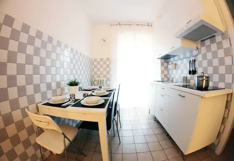 Compact, bright kitchen with a small dining table set for four; light grey checker tiles, white cabinets and natural light from a window create a clean, welcoming space.