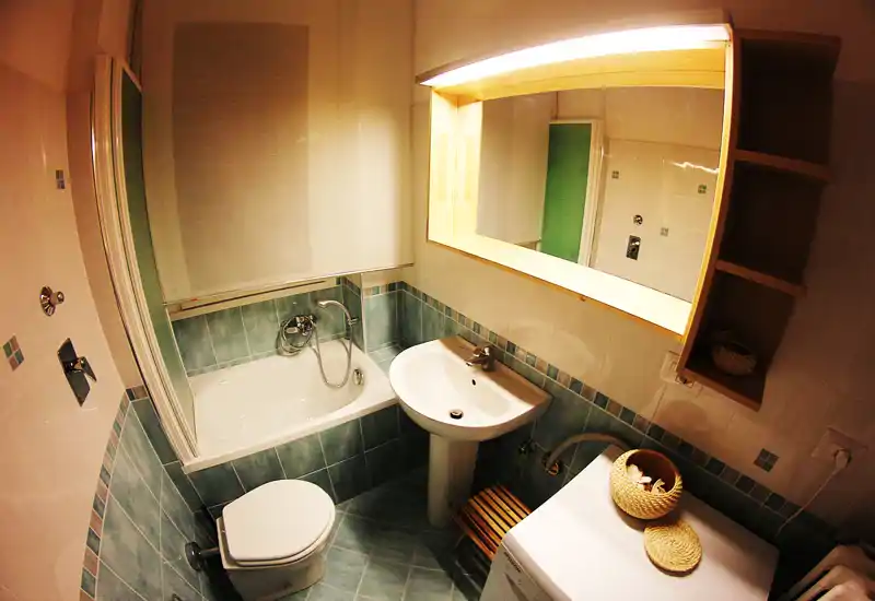 Baño compacto y bien iluminado con bañera, lavabo de pie, inodoro y espejo; azulejos verdeazulados en paredes y suelo para una apariencia fresca.