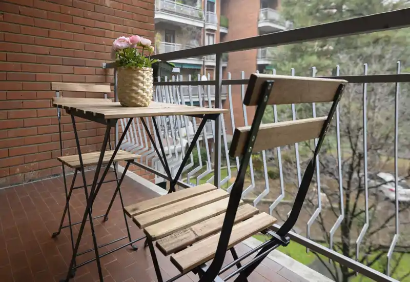 Balcone soleggiato con un piccolo set da bistrò in legno e un vaso decorativo con fiori, ideale per il caffè mattutino con vista sul cortile.