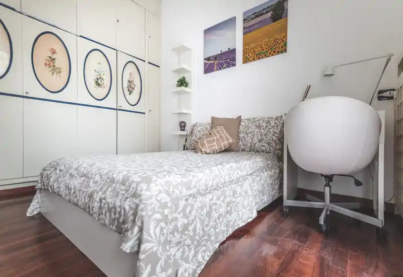 Chambre lumineuse et cosy avec un lit double à motifs, coussins décoratifs et placard intégré. Sol en bois chaleureux, tableaux muraux et chaise de bureau pour un petit espace de travail.