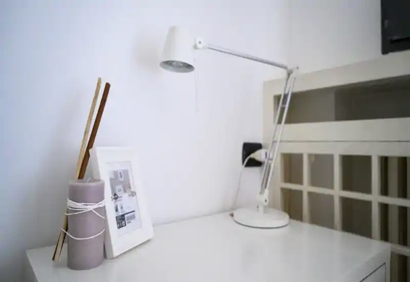 Coin de chambre minimaliste avec une table de chevet portant une lampe, une photo encadrée et une bougie décorative ; une partie du lit ou d’un lit superposé apparaît à droite.