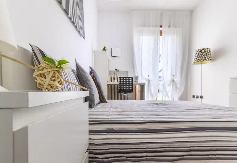 Dormitorio moderno y luminoso con cama doble bien hecha, cojines decorativos y mesita de noche. Cortinas translúcidas permiten la entrada de luz natural y un pequeño escritorio junto a la ventana aporta funcionalidad y calidez.