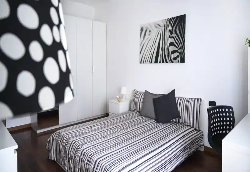 Dormitorio moderno y luminoso con ropa de cama a rayas, cojines coordinados y un cuadro de cebra sobre la cabecera. Mobiliario blanco y armario ofrecen almacenamiento; el escritorio y la lámpara aportan funcionalidad y estilo.