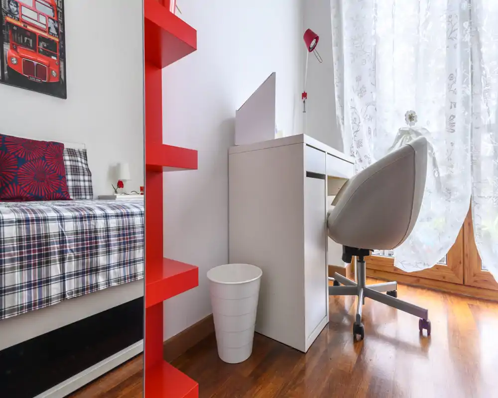 Rincón de dormitorio luminoso y moderno con cama visible con ropa a cuadros, un pequeño escritorio blanco y silla giratoria junto a la ventana soleada.