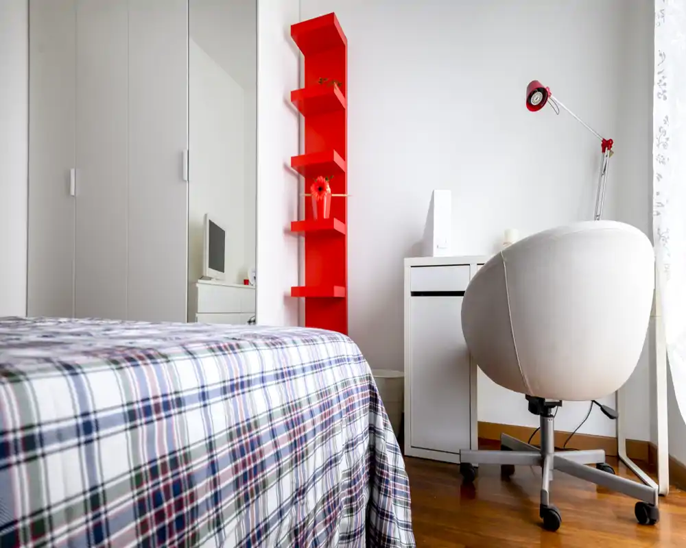 Dormitorio moderno y luminoso con colcha de cuadros en primer plano, un escritorio blanco y silla giratoria junto a la ventana, y una estantería vertical roja que aporta contraste de color.