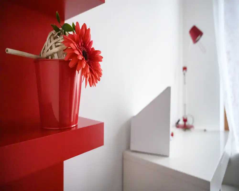 Plano cercano de un rincón de dormitorio moderno con acentos rojos: una flor roja en una maceta sobre una repisa y un escritorio blanco minimalista con una lámpara roja al fondo.