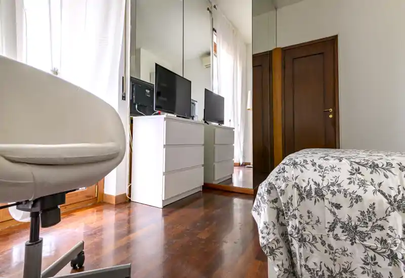 Bright, tidy bedroom with a patterned bedspread, mirrored wardrobe and a white dresser with TV—warm wooden floors and a comfortable chair by the window.