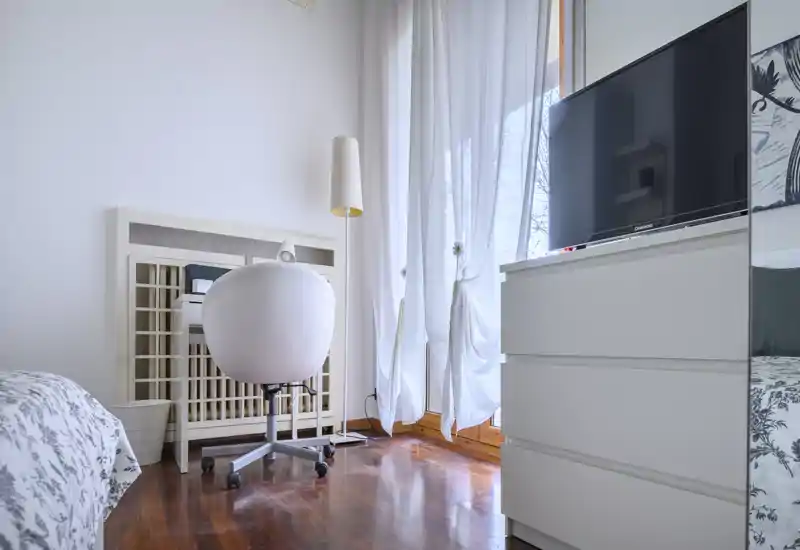 Bright, modern bedroom corner with patterned bedding, a rolling desk chair, tall sheer curtains and a dresser with TV — cozy and well lit for a comfortable stay.