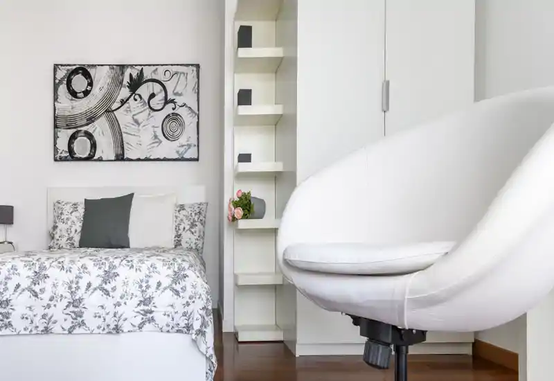 Bright, modern bedroom with a patterned duvet, decorative pillows and framed wall art; a comfortable swivel chair occupies the foreground creating a stylish, lived-in feel.
