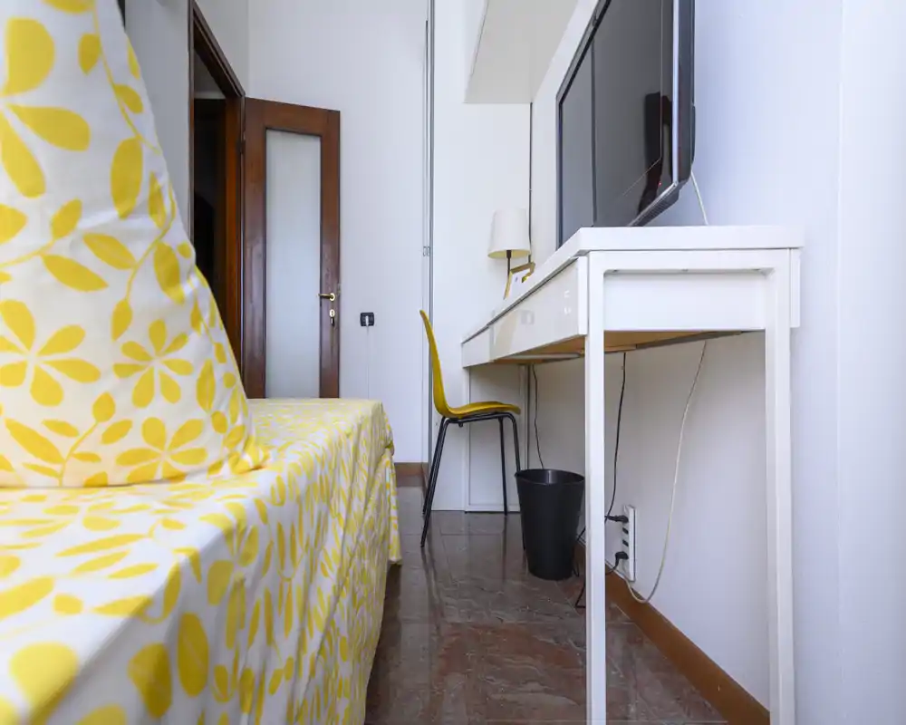 Camera compatta e luminosa con copriletto giallo a fantasia, una scrivania con TV sopra e una sedia — perfetta per un ospite singolo o soggiorni brevi.