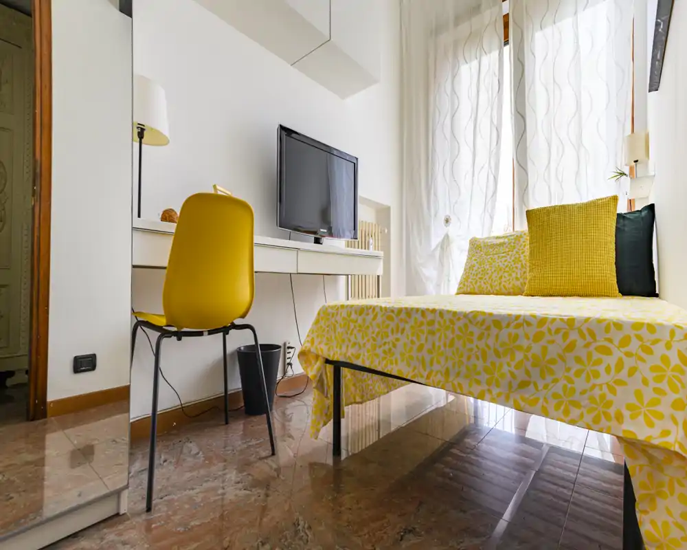 Camera da letto luminosa e moderna con copriletto floreale giallo, cuscini coordinati e una scrivania con TV — ideale per un soggiorno singolo o doppio allegro.