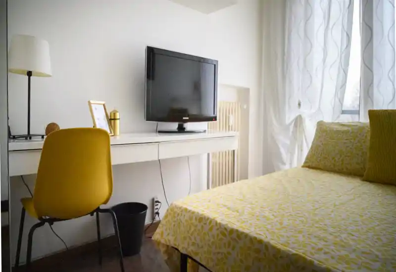 Bright, cozy bedroom with a yellow patterned bedspread, matching cushions and a desk with a TV. Large window with sheer curtains provides natural light and a warm, welcoming atmosphere.