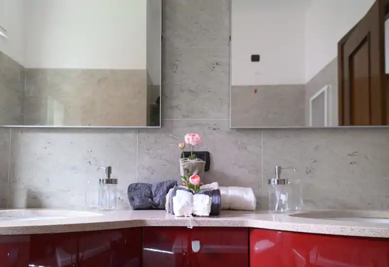 Mueble de baño moderno y luminoso con doble lavabo, armarios lacados en rojo, dos espejos y toallas dobladas con un pequeño arreglo floral.