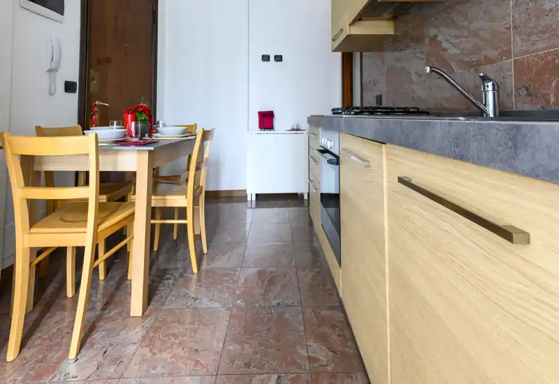 Well-lit modern kitchen with wooden cabinets, integrated oven, sink and a small dining table set for four — compact and functional for everyday meals.