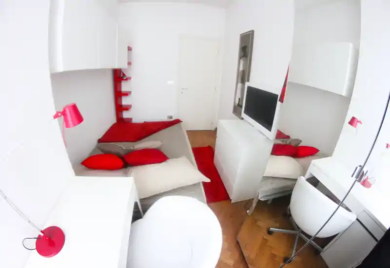 Compact modern bedroom with white furniture and bold red accents: bed with multiple pillows, a dresser with TV, desk and chair. Bright, stylish and cozy space ideal for a single occupant.