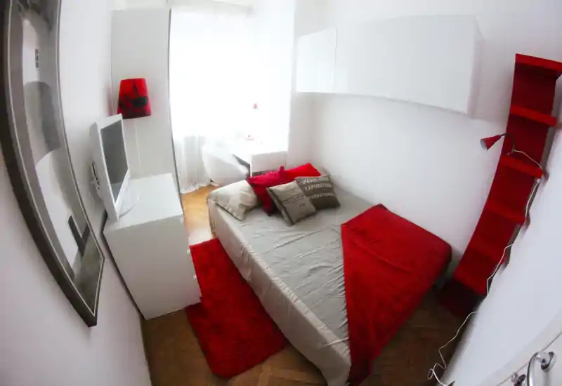 Compact, stylish single bedroom with a neatly made bed, red accents, wall cabinets and a bright work corner by the window.